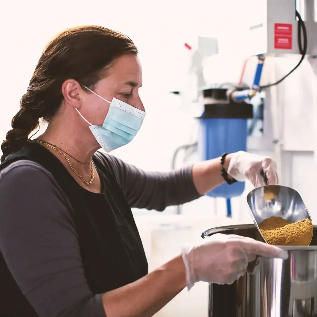 Person in a lab setting wearing gloves and a mask, handling a container with a yellow substance.